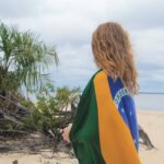 En kvinde med det brasilianske flag om skuldrene kigger ud over floden fra en sandbred